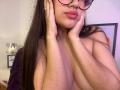 IrisBlair with long brown hair, wearing glasses, hands on cheeks, pink lips, and visible large bare boobs, slightly covering her nipples with her fingers.