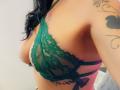 FrenchShanna in green lace bra, showing her tits, tattoo on right arm, black curly hair, light brown skin, side view, indoors, sexy, lingerie, partial tattoo visible.