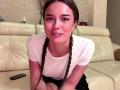 DrusillaMaiava, with braided brown hair, white t-shirt, and black skirt, sitting on a cream leather sofa, smiling, showing small tits, and a cute face, with a phone nearby.