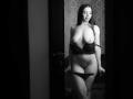 Black-and-white photo of CelesteNoire with long dark hair, standing, topless, wearing a black bra, lowered black panties, showing her tits, ass, and pussy. She's smiling softly.
