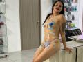 AriannaSeete with painted blue handprints on her tits, belly, and ass, wearing white panties, leaning on a counter in a room with shelves of products. She's smiling.