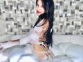 EstrellaXHot with wet black hair, smiling, sitting in a bubbly bathtub, showing her ass, with soapy water covering her back and buttocks. She's wearing a thong.