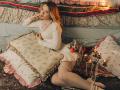 KylaHemeke with blonde hair, wearing a white dress and pearls, sitting on a rug with floral pillows, legs spread, candles and roses, looking seductively at camera.