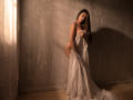 KarlaSoto with long brown hair, holding a white, sheer cloth over her tits and ass, standing in a dimly lit room, shadows on the gray wall.