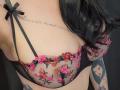 De zwarte kanten beha van FrenchShanna, met roze bloemenborduurwerk, laat haar borsten zien. Ze heeft zwarte bandjes en een klein zwart strikje. Ze heeft tatoeages op haar arm en borst..