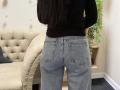 Dalaila's ass in light blue jeans with pockets, standing with her back to the camera, wearing a black long-sleeve top, in a room with a beige tufted chair.
