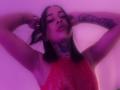 HollyCattaleiya with arms raised, wearing a red, sparkly top, showing tattoos on her neck and arm, under purple light, looking relaxed and confident.