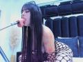 AlisonTatcher with long black hair, tongue out, sucking a microphone, wearing fishnet dress, showing tattoos on right arm, bent over, blue-lit room, black headband, pink tongue.