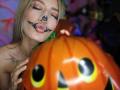 Kaiix met een zwart hartje op haar neus, samengeknepen lippen en blond haar, die een grote oranje pompoen vasthoudt met zwarte ogen en gele pupillen, en waarbij haar kleine borsten zichtbaar zijn..