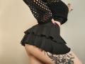 Niferttiti's ass in a black, ruffled skirt, wearing a black, fishnet top, with a large floral tattoo on her right thigh, hand against a wall.