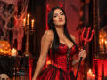 AriiaMon with red devil horns, black hair, red corset, and large tits, holding a red trident, surrounded by candles and Halloween decor, looking sexy and confident.
