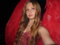 AkilahGhil with long, wavy blonde hair, pink eyeshadow, and a pink sequined dress, showing her tits slightly, wearing red wings in a dark, blurry background.