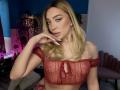 JessicaClarks with blonde hair, wearing a sheer red crop top, showing off her tits, hand near her face, pink nail polish, pearl earrings, and a sexy, confident look.