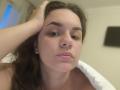Dreia has puffy pink lips, brown eyes, and a hand in her dark brown hair, with a neutral expression, and is topless, showing her bare shoulders and a white pillow.