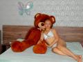 Anshante with small tits and a slim ass, wearing white lingerie, sits on a bed next to a giant brown teddy bear, in a room with patterned wallpaper.