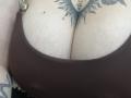 DiLaria's large tits in a brown bra, showing a sunburst tattoo between her cleavage and a small black dot tattoo above. She's also wearing a gold button.
