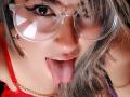 AmiiRosse with tongue out, wearing clear glasses and red top, has wet tongue and slightly open mouth, gray hair, and gold necklace, close-up shot.