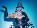 Une femme avec du maquillage bleu sur le visage, un haut-de-forme noir et un corset, portant des gants noirs et une volante blanche, touchant son menton, avec des tatouages sur le bras droit, des motifs floraux et des roses roses..