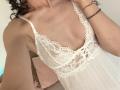A lingerie branca de renda da SweetClaraa, com um decote profundo, que mostra seios pequenos e um corpo esguio e bronzeado. Seus cabelos cacheados e a pulseira no pulso são visíveis..
