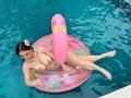 MaddyCarina with dark hair, pink flower in hair, lying on pink inflatable ring in pool, naked, large tits, tattoos, legs crossed, holding ring, pink inflatable ring with yellow flowers.