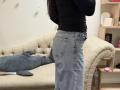 Dalaila stands in front of a beige tufted couch, wearing a black top and blue jeans, with her back to the camera, showing her ass and back pocket.