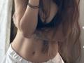 VelvetRay's small tits, covered by a sheer top, with long brown hair, white pants, and a black bracelet, holding her arms up, showing a slim, flat-stomach.