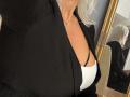 Hotsexydolly with gray hair, wearing black blazer, white bra, and black strappy bra, showing her tits in mirror reflection. Gold necklace with cross. Soft lighting.