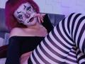 ViolettClay with red hair, white face paint, black lipstick, and striped thigh-highs, wearing a black off-shoulder top, resting her chin on her hand. Boobs partially visible.