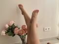 MysteryLaddy's bare legs are crossed at the ankles, with pink-painted toes, near a vase of pink roses on a white tile floor. Two electrical outlets are visible.