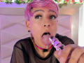DannyRouds with pink hair, black mesh top, pink choker, tattooed arm, and earrings, smiling, hand on head, in a softly lit room with floral lights.