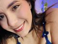 LucieMilner with a cute smile, wearing blue lace lingerie with flower details, black choker, and pink lipstick. Her brown eyes and long hair are prominent. Boobs are visible.