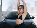 Milagres with arms raised, wearing a black bikini top, black high-waisted pants, and sunglasses, standing in front of a black car with water spraying. She has a tattoo.
