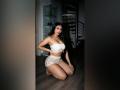 MiilaWx kneels on wooden floor in white lace bra and panties, showcasing her big tits and curvy ass, with long black hair and red lipstick in dimly lit room.