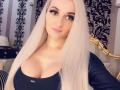 Mademoiselle Bella, con lunghi capelli biondo platino, occhi azzurri, rossetto rosa, top nero che mette in risalto un seno generoso, una testiera dorata e tende nere a motivi in evidenza sullo sfondo..