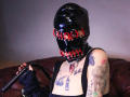 AmySpider with black latex mask, red eyelets, holding a black whip, tattoos on chest and arm, wearing black tank top, seated on brown leather couch.