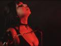 MadisonHetfield with red-lit face, black hair, eyes closed, head tilted back, wearing black choker, deep cleavage, holding chain in front, dark background.