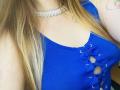 A blusa azul canelada da KarKutie, com amarração na frente, que mostra o decote, e um colar de prata. Cabelo loiro comprido, tatuagem no ombro direito..