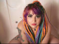 GineBralkino with colorful dreadlocks, purple and orange hair, tattoos on her arm, and a necklace, looking up with eyeliner and red lipstick, in a white, slightly blurred background.