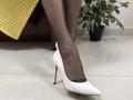 Rorria's black pantyhose-clad legs with white pointed high heels on a shiny tile floor, yellow ribbed fabric in the background, and a green potted plant to the right.