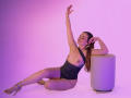 AnabelleRoss with tits exposed, wearing a black one-piece, sitting on the floor, arms raised, legs crossed, high heels, leaning on a white cylinder, pink-purple lighting.