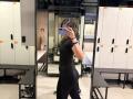 HeikeCarchi in a black tight dress and leggings, showing a curvy ass and toned arms, taking a mirror selfie in a gym with white lockers.