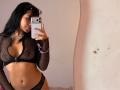 VannesaSaenz, a woman with medium brown skin and long black hair, takes a mirror selfie. She wears a black mesh long-sleeve top with a deep V-neck, exposing her cleavage, and matching black mesh panties. She has a slim, toned physique. She holds a white iPhone in her right hand, with a silver necklace and purple beaded bracelet on her left wrist. The wall behind her is plain and slightly textured.