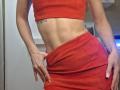 Close-up photo of MilaYanis with a slim, toned midriff, wearing a red crop top and matching red skirt. She's pulling the skirt down, exposing her hip and hand with painted nails.