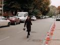 VirgieCrotts with brown coat, black hat, and boots, walking on a street, holding a small brown purse, with parked cars and orange traffic cones in the foreground.
