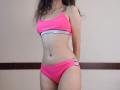 VikkiRi with pink bra and panties, slim with small tits and a flat ass, standing, arms behind her back, long wavy brown hair, light skin, in a simple room.