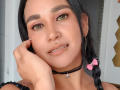 EdenIvyLm a un visage mignon avec des cheveux foncés, des yeux verts, du fard à paupières rose, un anneau au nez, un piercing aux lèvres, un collier noir, un nœud rose et des boucles d'oreilles cerceaux, et elle touche sa joue..