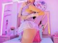 MelisaDaltonn in purple lace bra, white sheer skirt, and gloves, biting finger, showing her ass, standing in a pink-lit room with a clock and mirror in the background.
