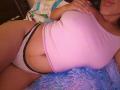 AntoniaBrunni lying on a bed, wearing a pink tank top that shows her big boobs and white polka dot panties with black trim. She has a fluffy blue pillow.
