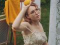 TeresaBirt with glasses, holding a lemon over her head, wearing a floral dress with ruffled straps, yellow cloth draped on a chair, white lace curtain.
