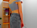 Steisii has a cute, slim ass in denim jeans, wearing a gray off-shoulder crop top, standing next to a wooden shelf with a purple plant and bottle.